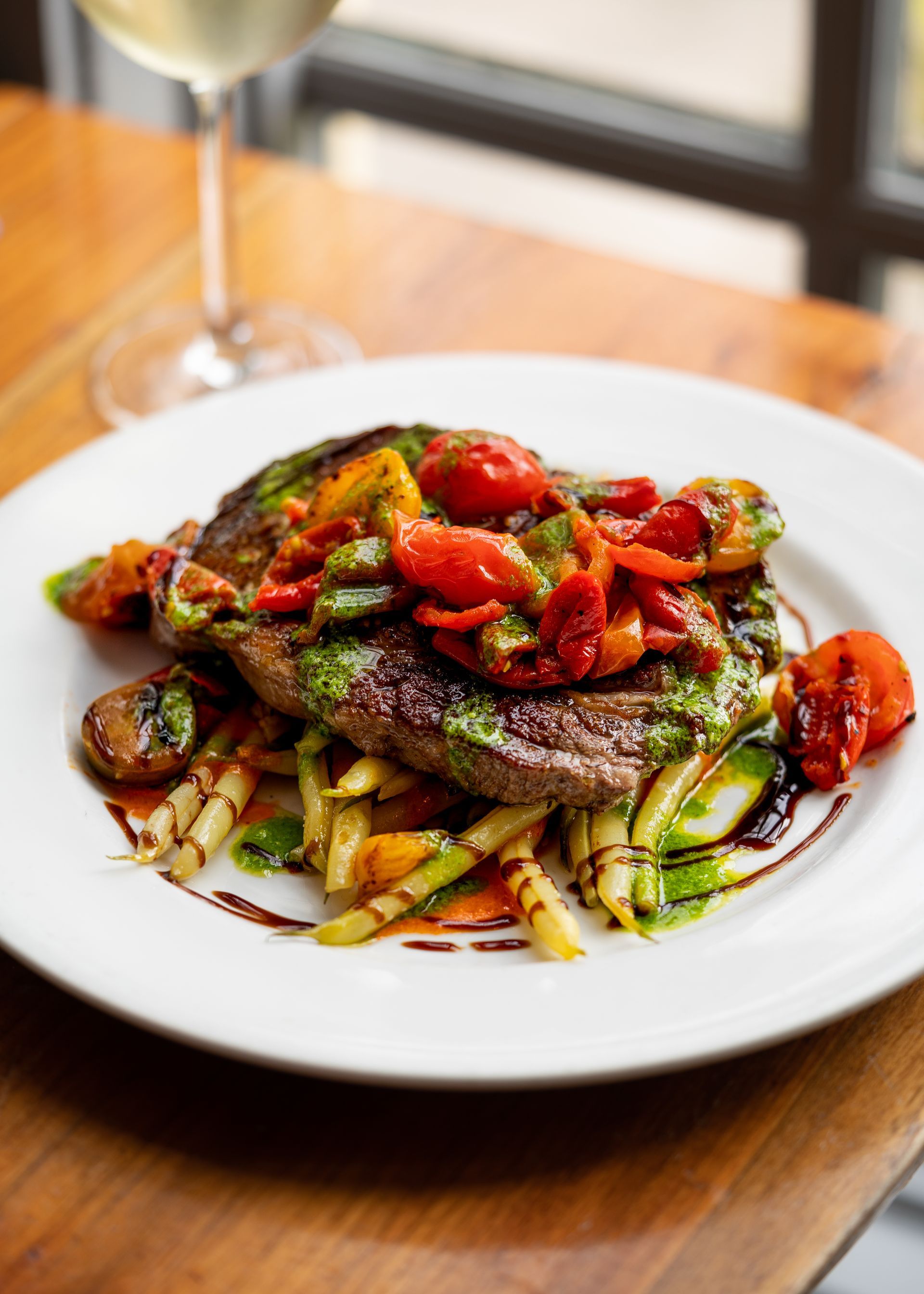 A white plate topped with meat and vegetables next to a glass of wine.