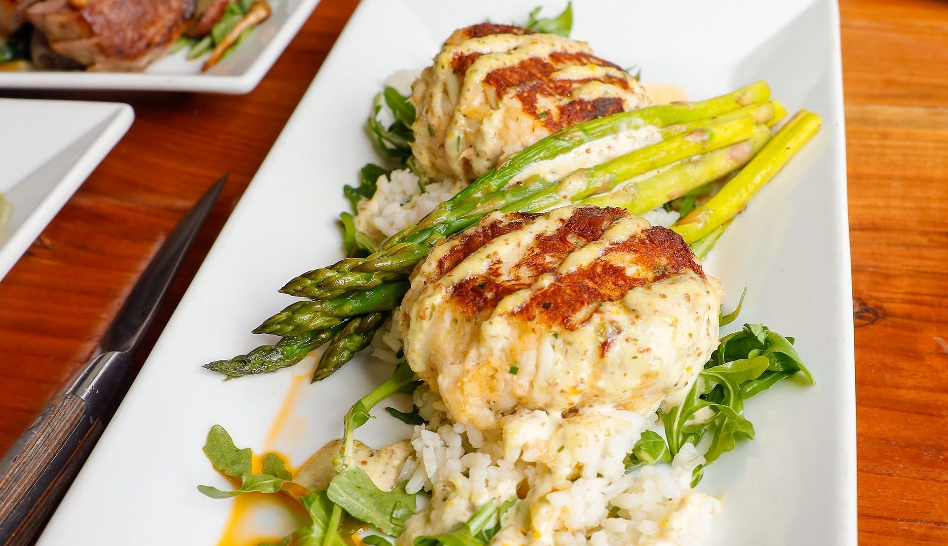 Crab cakes with grilled asparagus and rice on a white plate, set on a wooden table.