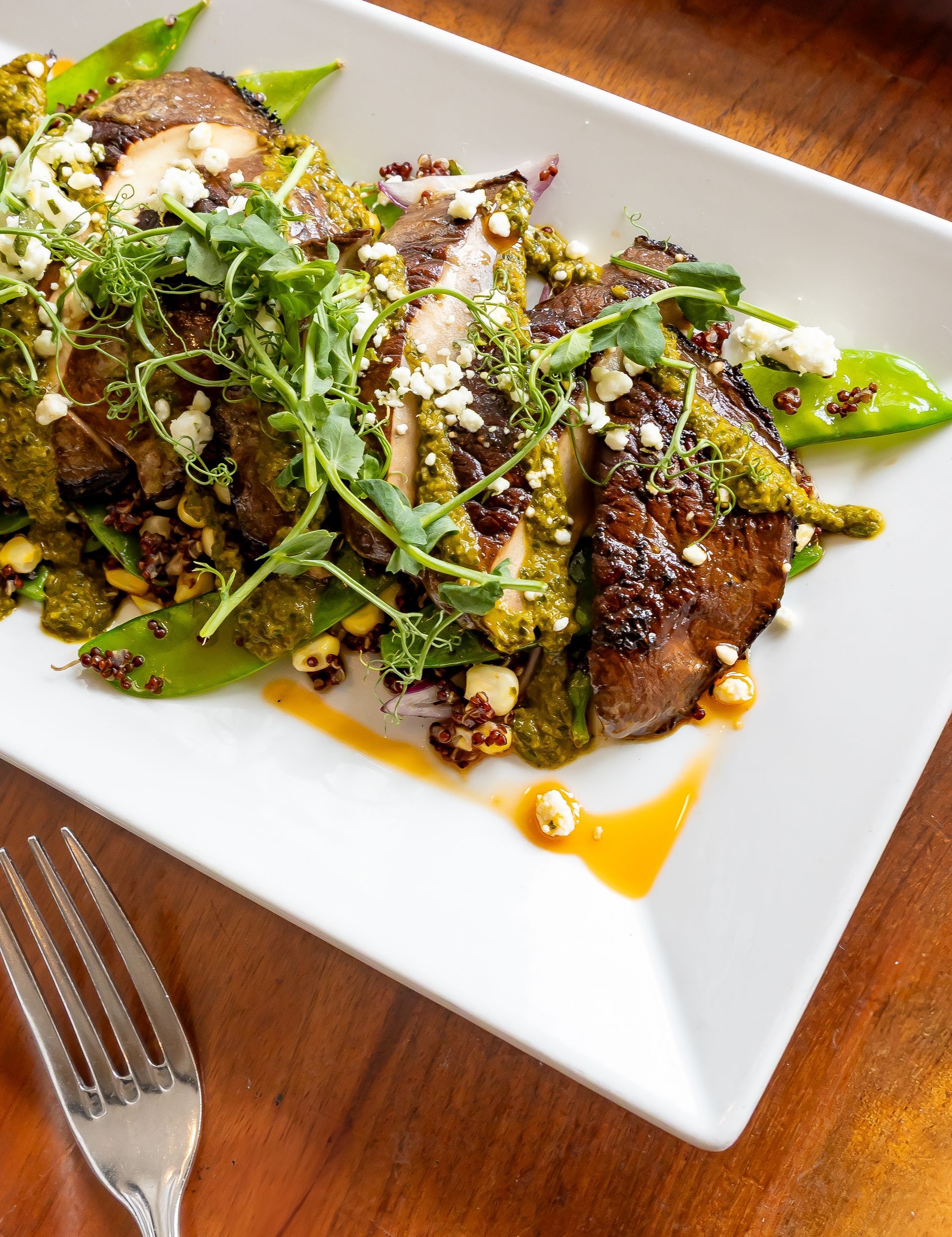 A white plate topped with meat and vegetables next to a fork on a wooden table.