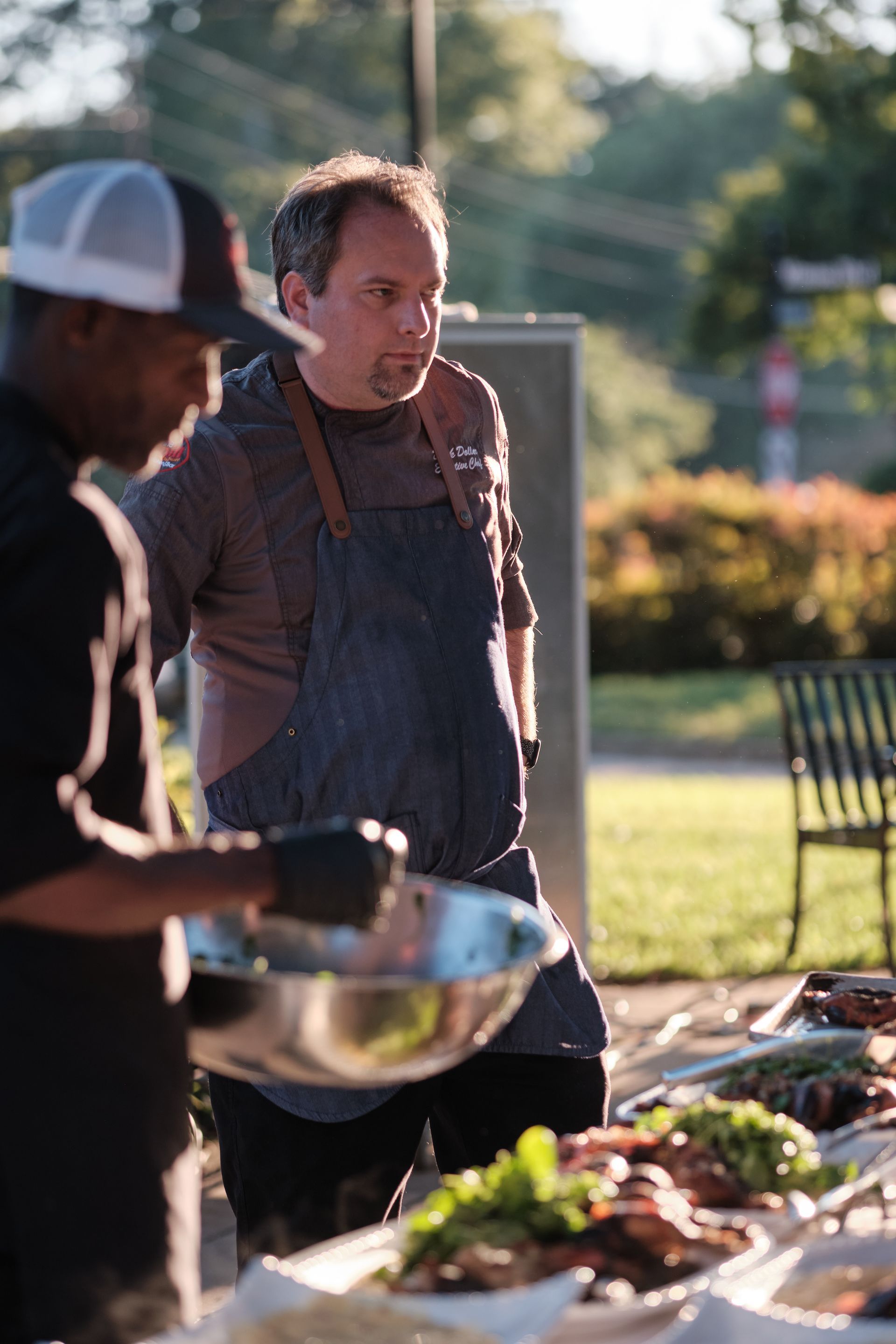Two chefs at a food station. One holds a bowl, the other in an apron observes the food with a thoughtful expression.