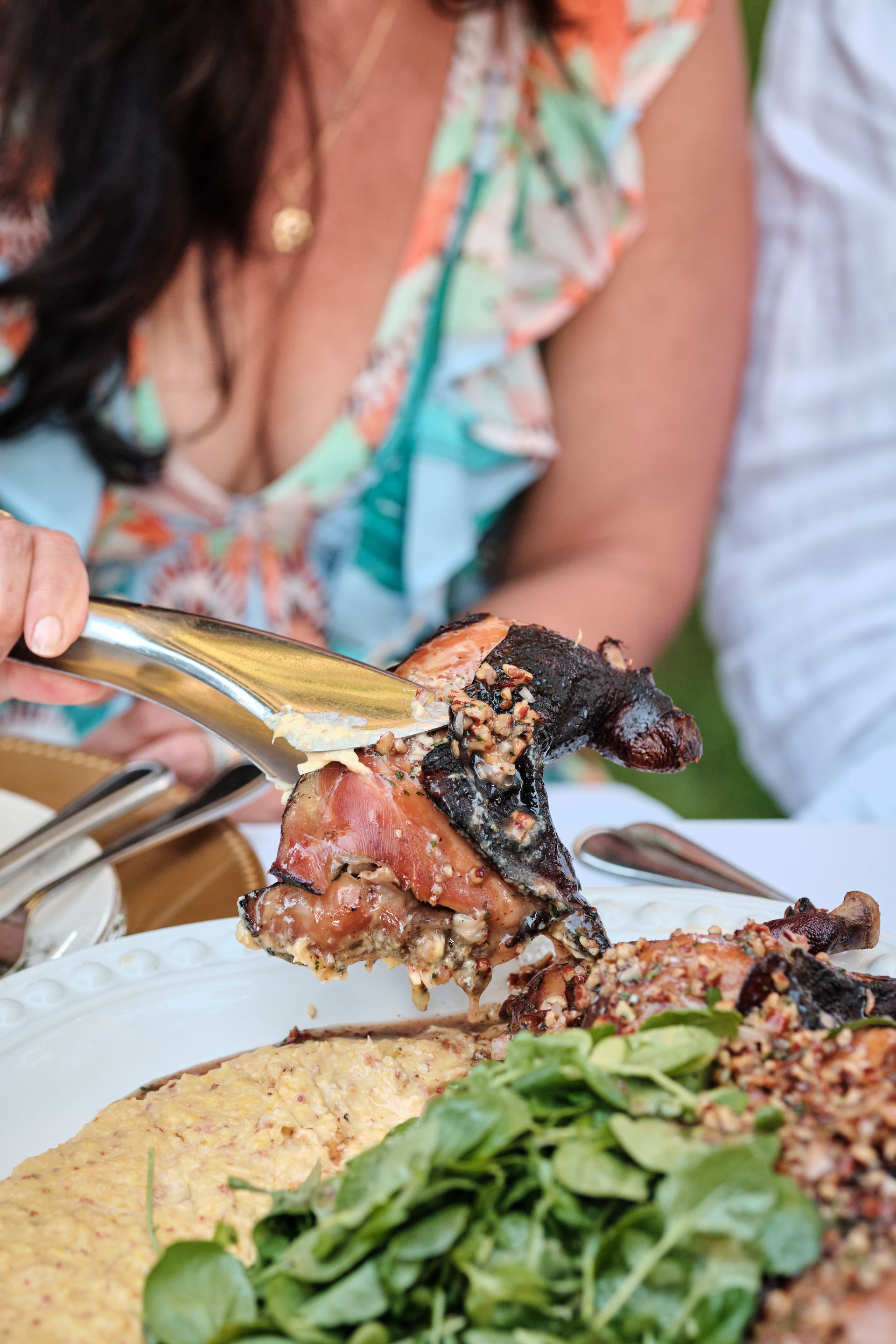 Woman using tongs to serve roasted meat from a platter; greens and a creamy sauce are visible.