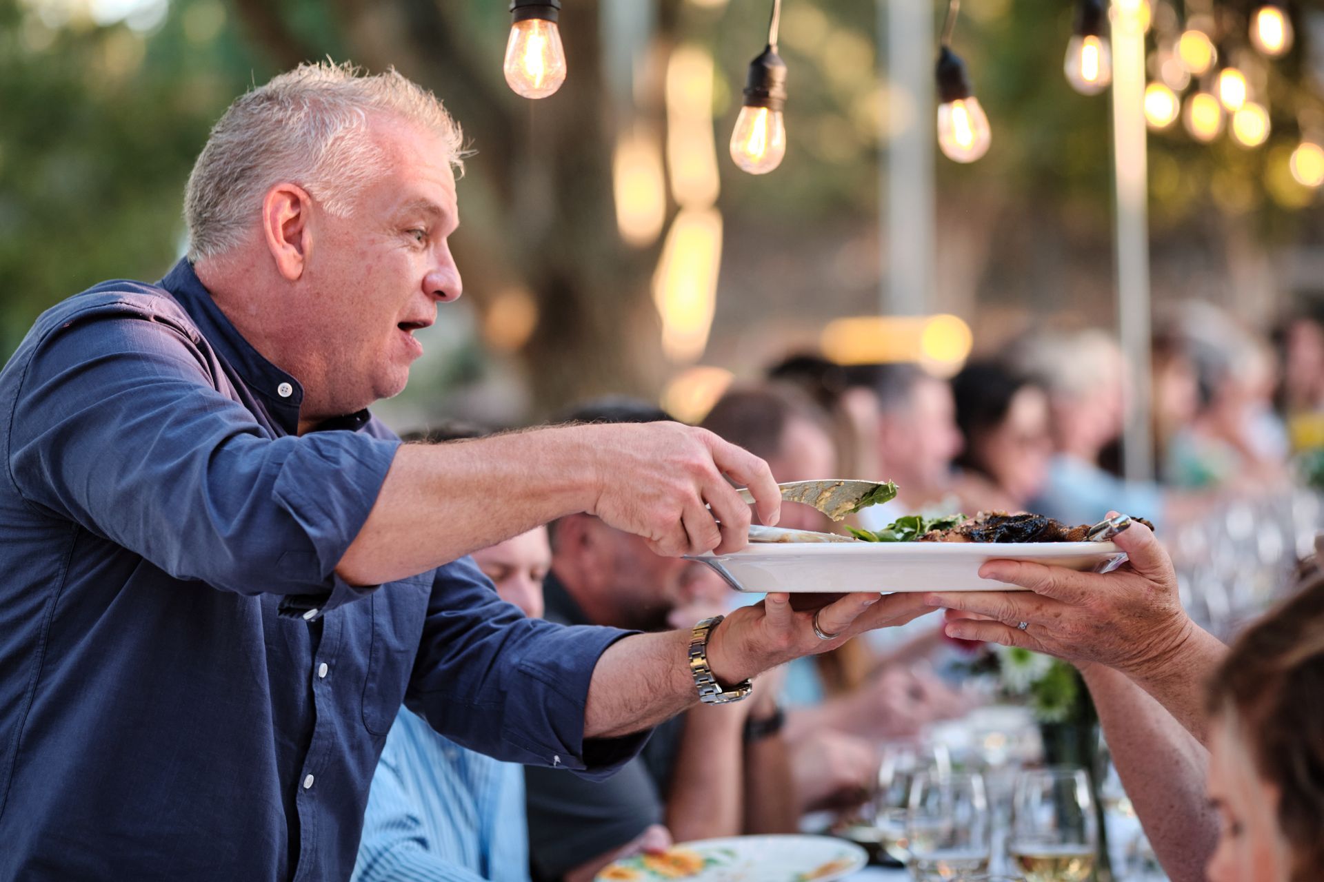 Man in blue shirt serving food on a white plate at an outdoor dinner party.