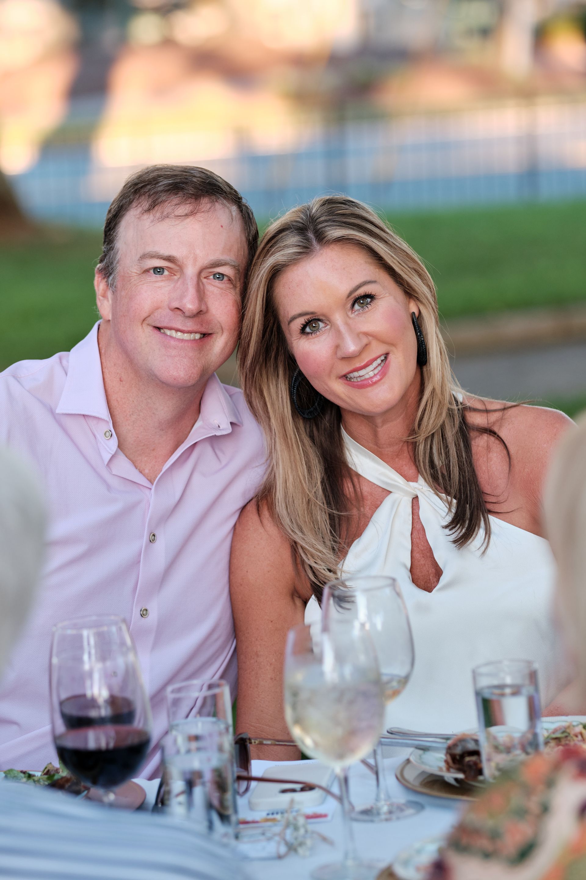 Man and woman smiling, at a table. Man in pink shirt, woman in white dress. Outdoors, blurry background.