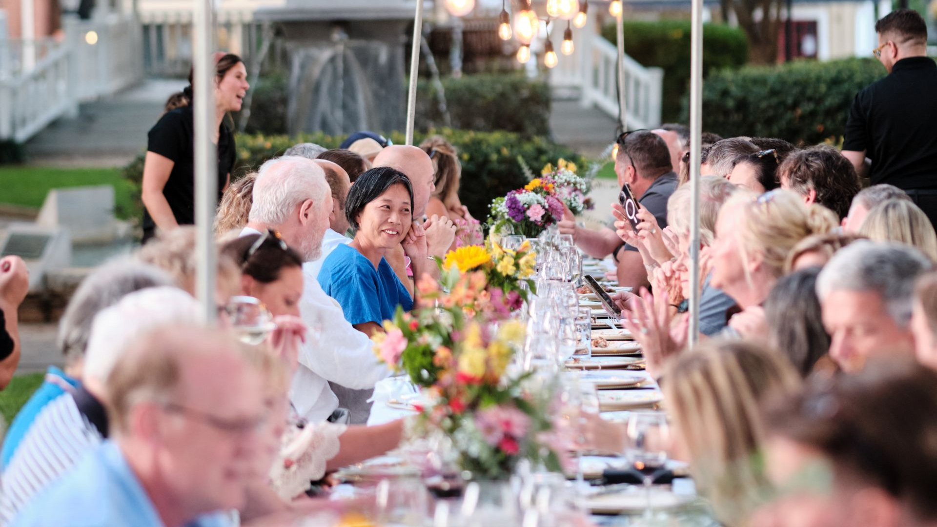 A group of people are sitting at long tables in a park.