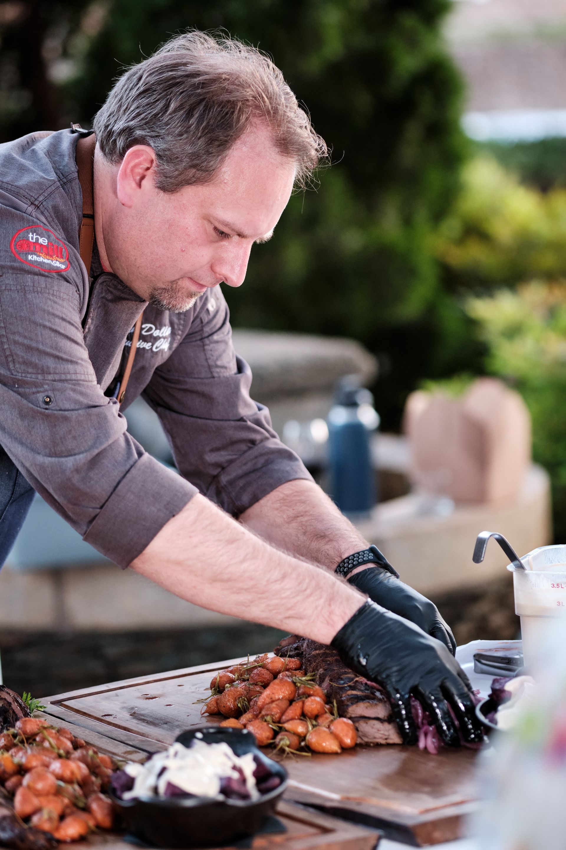 Chef cutting meat with gloved hands outdoors. Carrots and other food items are on the table.