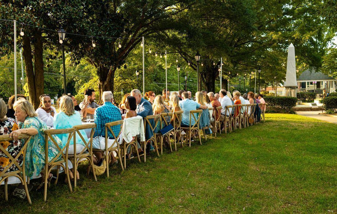 A group of people are sitting at long tables in a park.