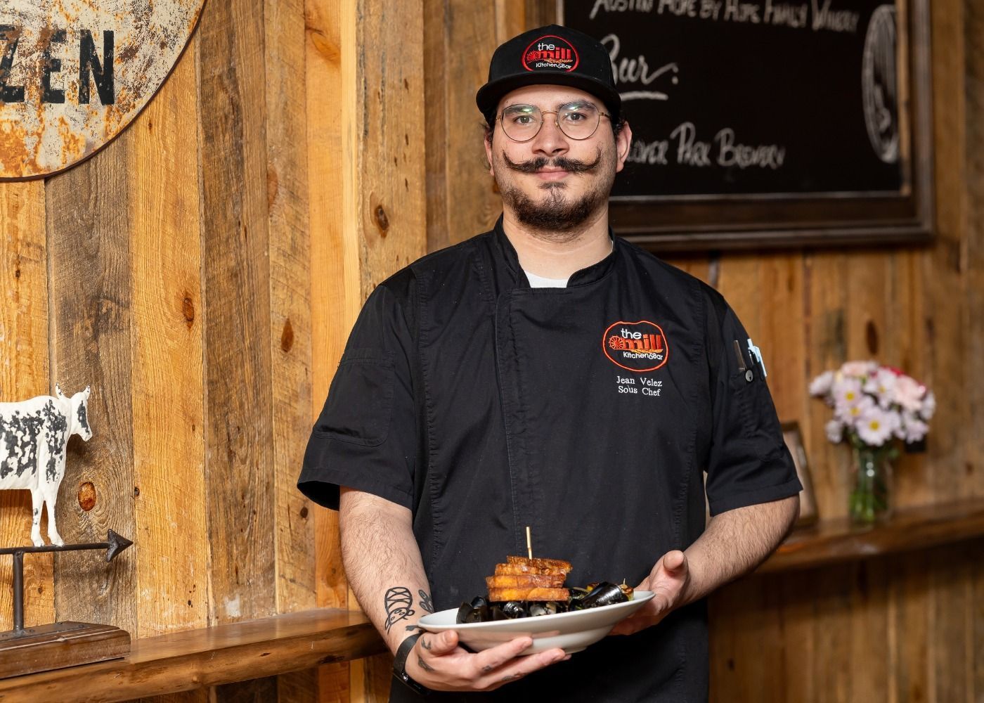 A chef is holding a plate of food in front of a wooden wall.