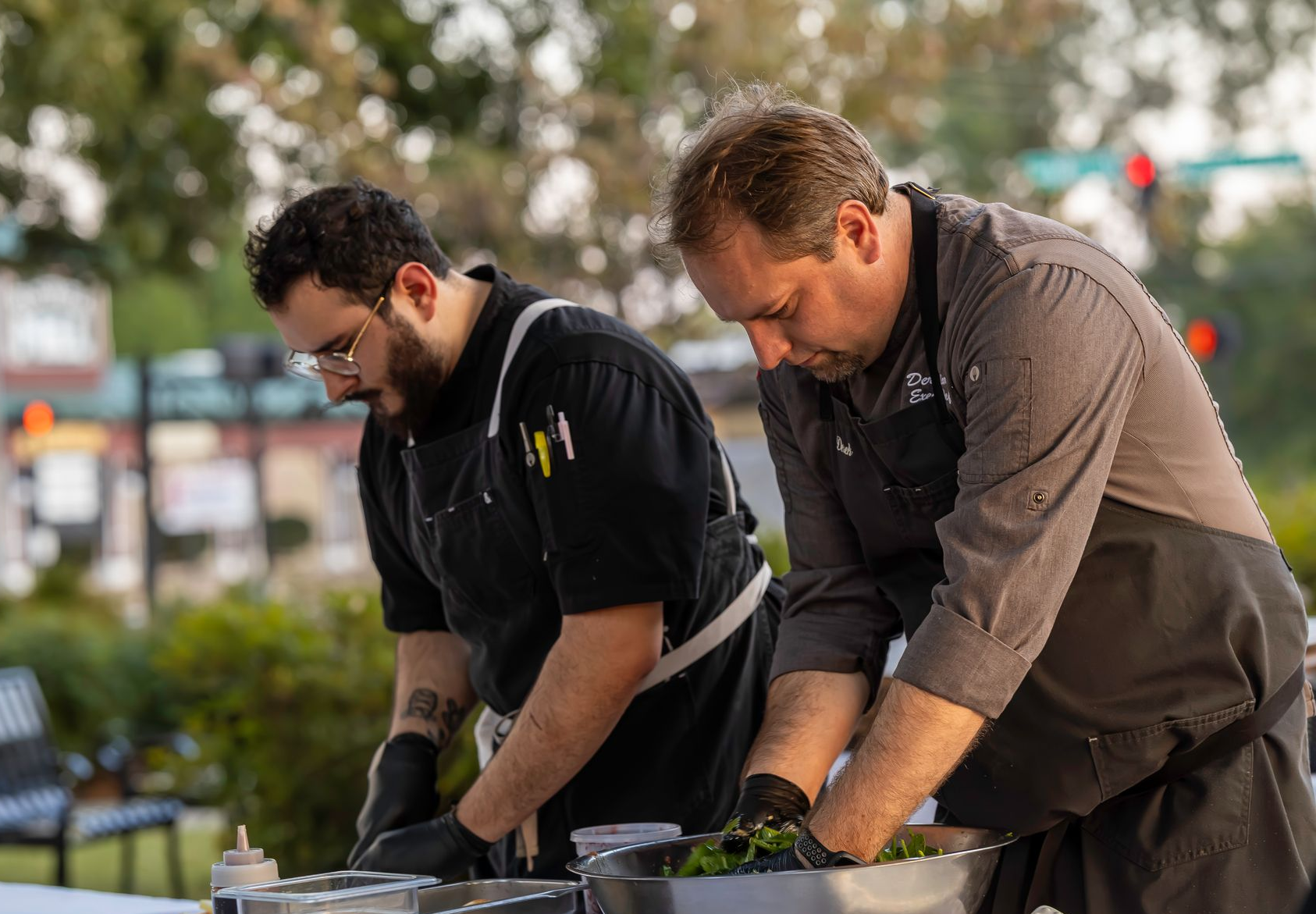 Two chefs in black uniforms, working outside at a food event, mixing ingredients in bowls.