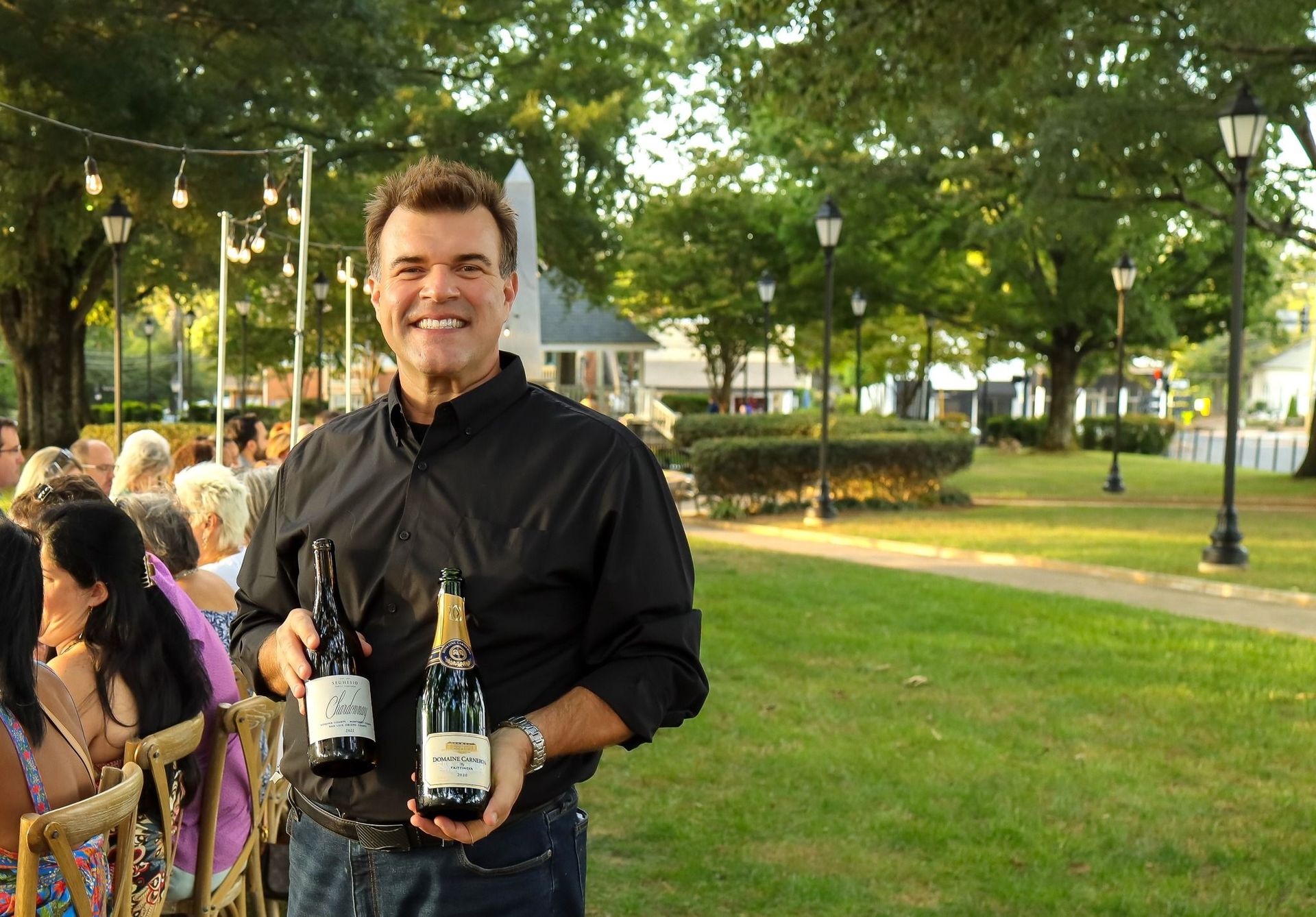 A man in a black shirt is holding two bottles of wine.