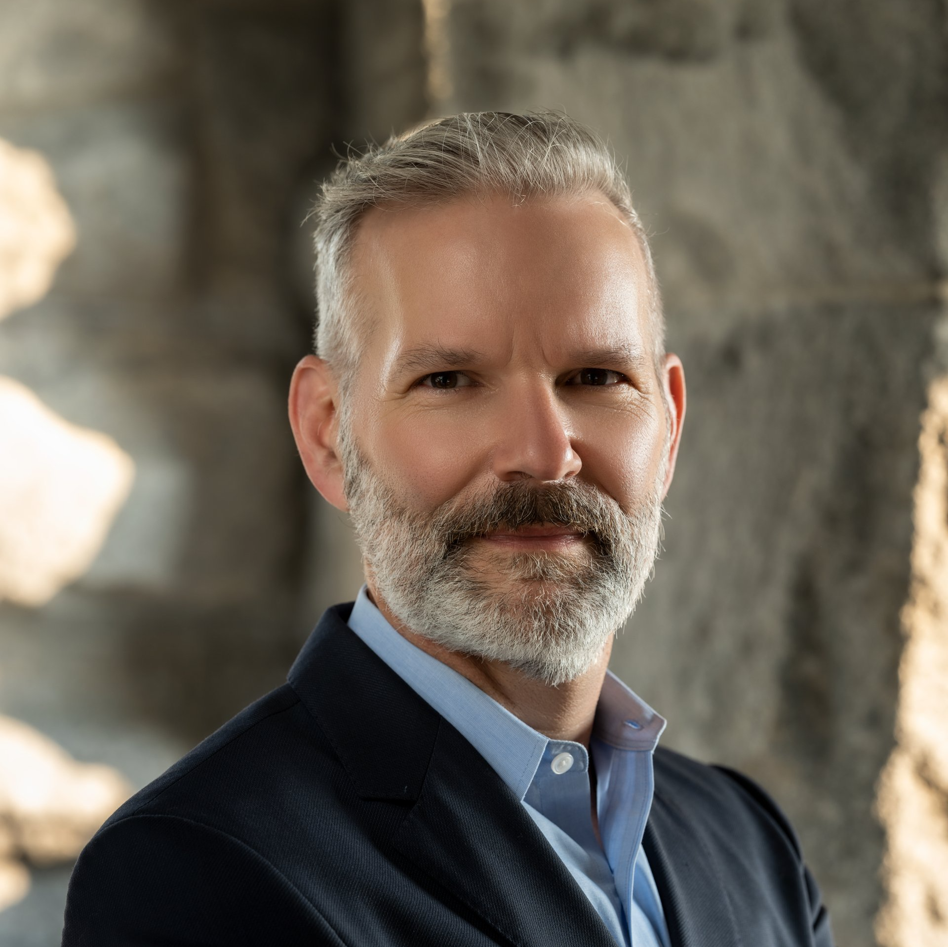 A man with a beard is wearing a suit and a blue shirt.