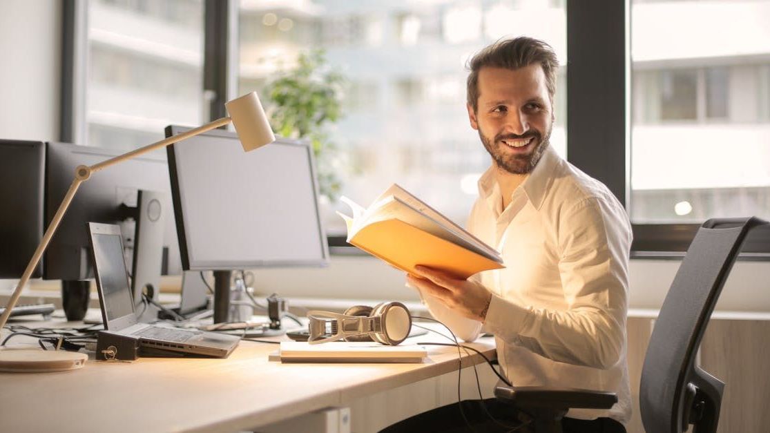 A man is sitting at a desk in an office holding a book.