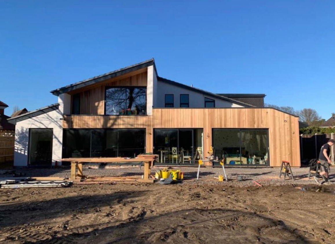 Modern house under construction with wood siding, large windows, and a person working outside on a sunny day.