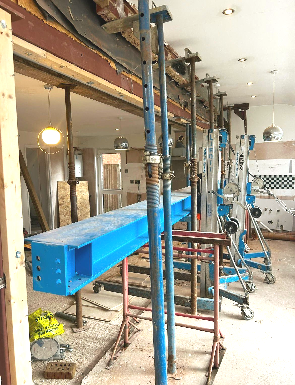 Interior construction site with blue support beams, metal scaffolding, and exposed wooden framing.