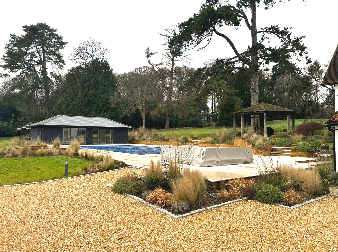 Exterior view of a pool area with a covered pool, landscaping, and a small building under an overcast sky.