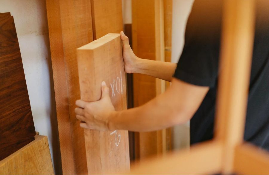 Person holding a light-colored wooden plank in a wood shop, surrounded by stacked lumber.