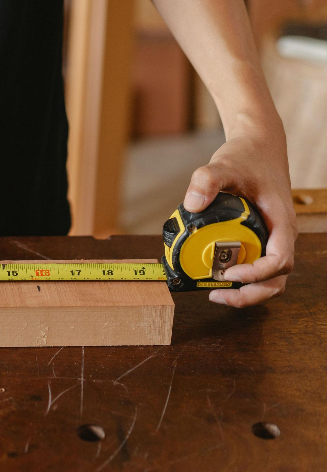 Hand measuring a wooden block with a yellow tape measure on a workbench.