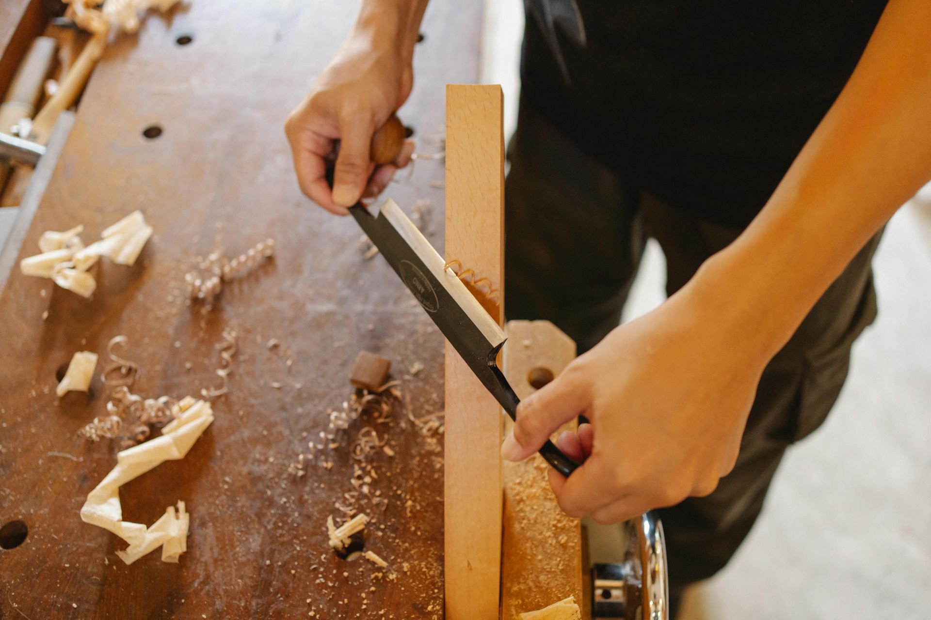 Person using a drawknife to shape a wooden board on a workbench, wood shavings present.