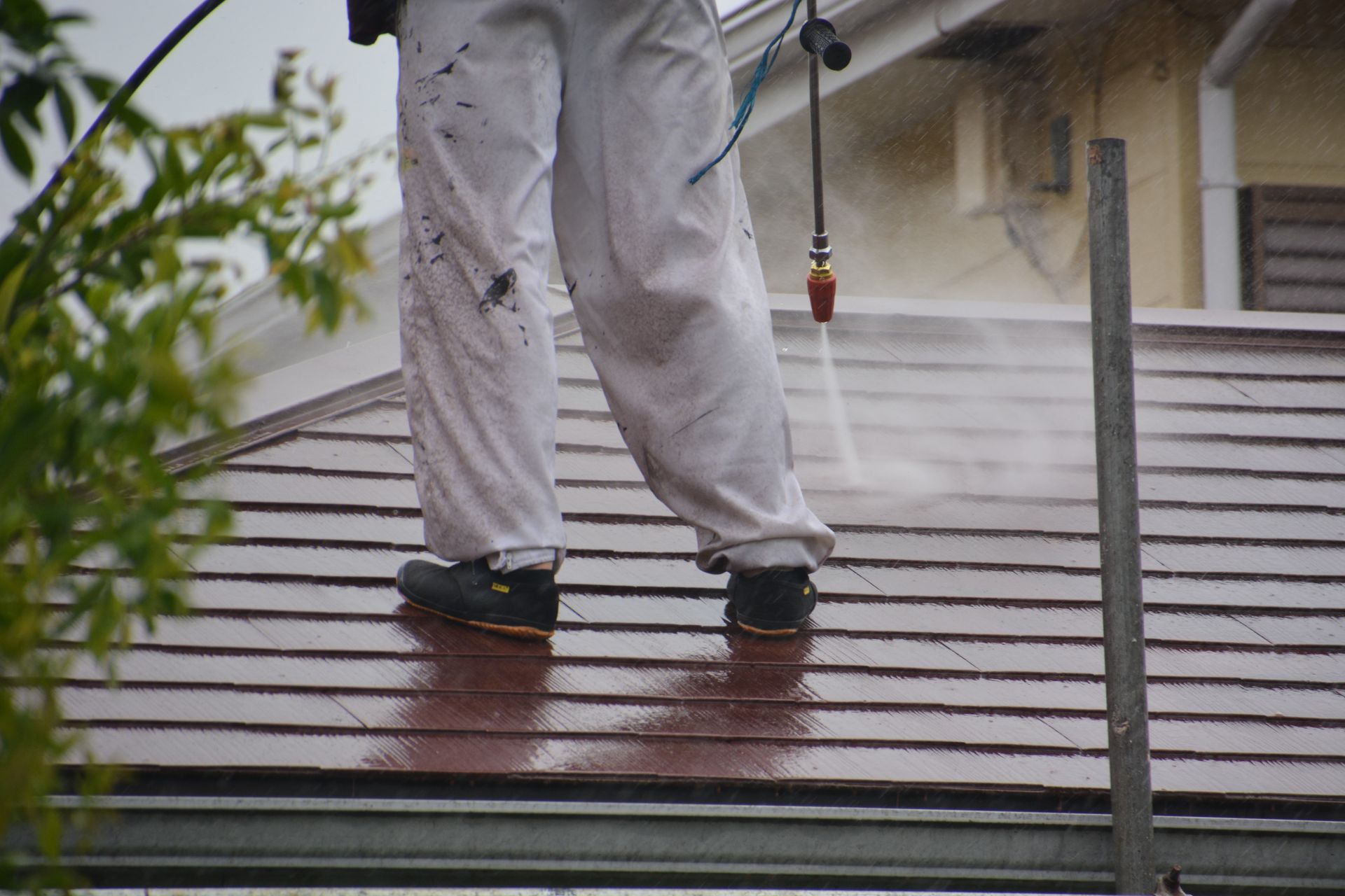 Person in white overalls power washing a brown shingled roof, steam visible, in a residential setting.