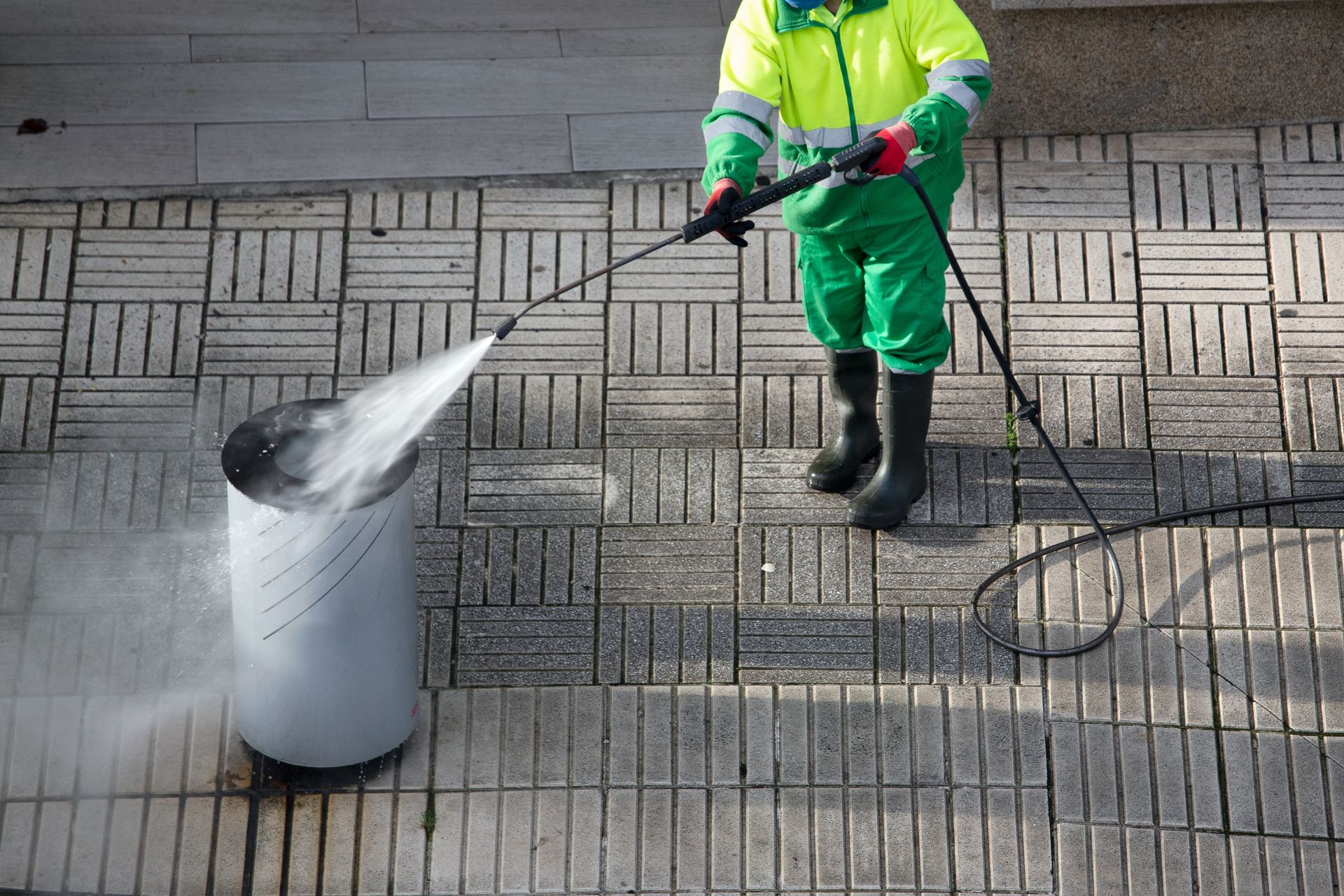 Person in green work suit power washing a cylindrical trash can on a brick walkway.