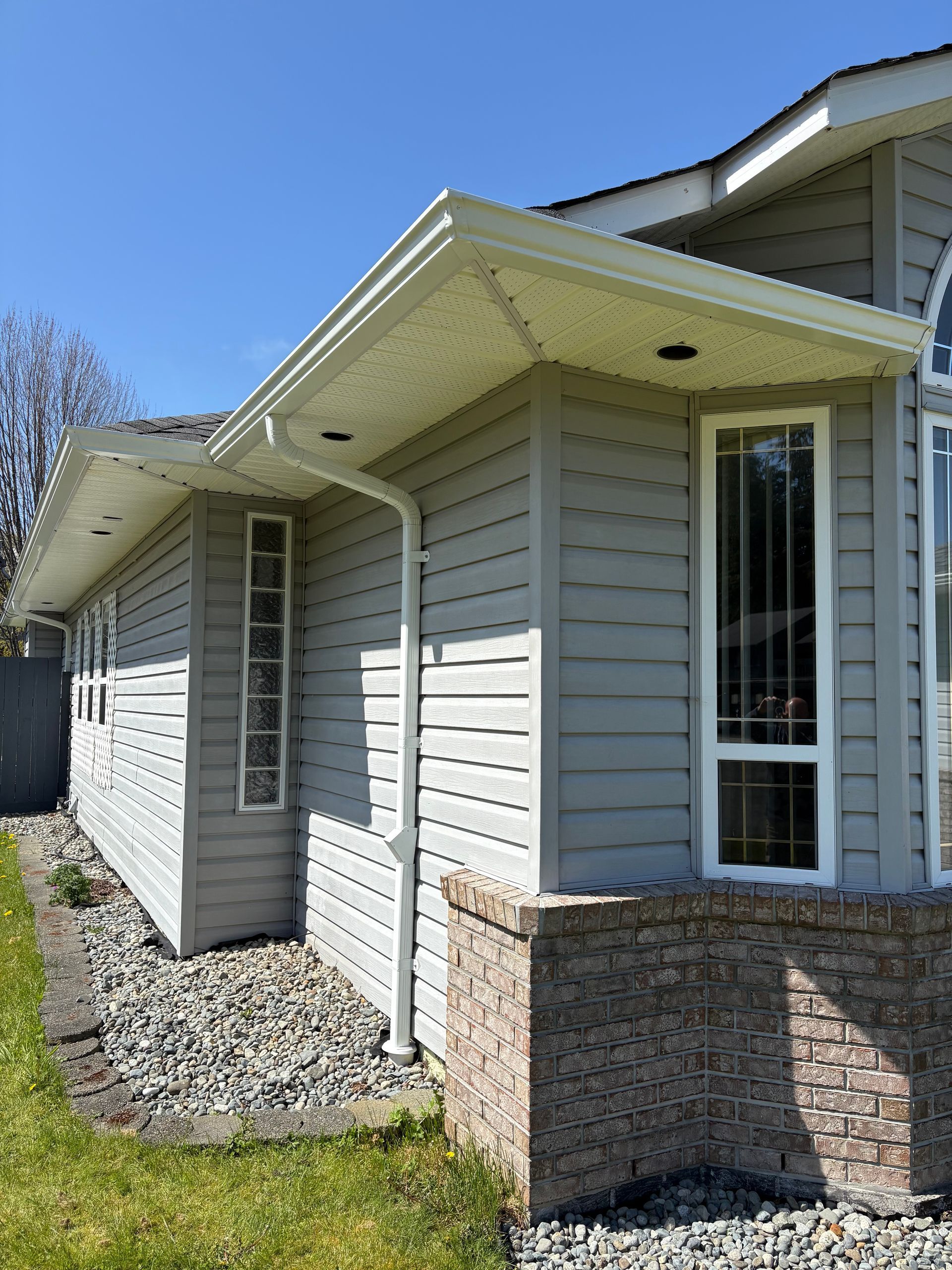 Gray house exterior with white trim, gutters, and downspout; brick base, rocky landscaping, blue sky.