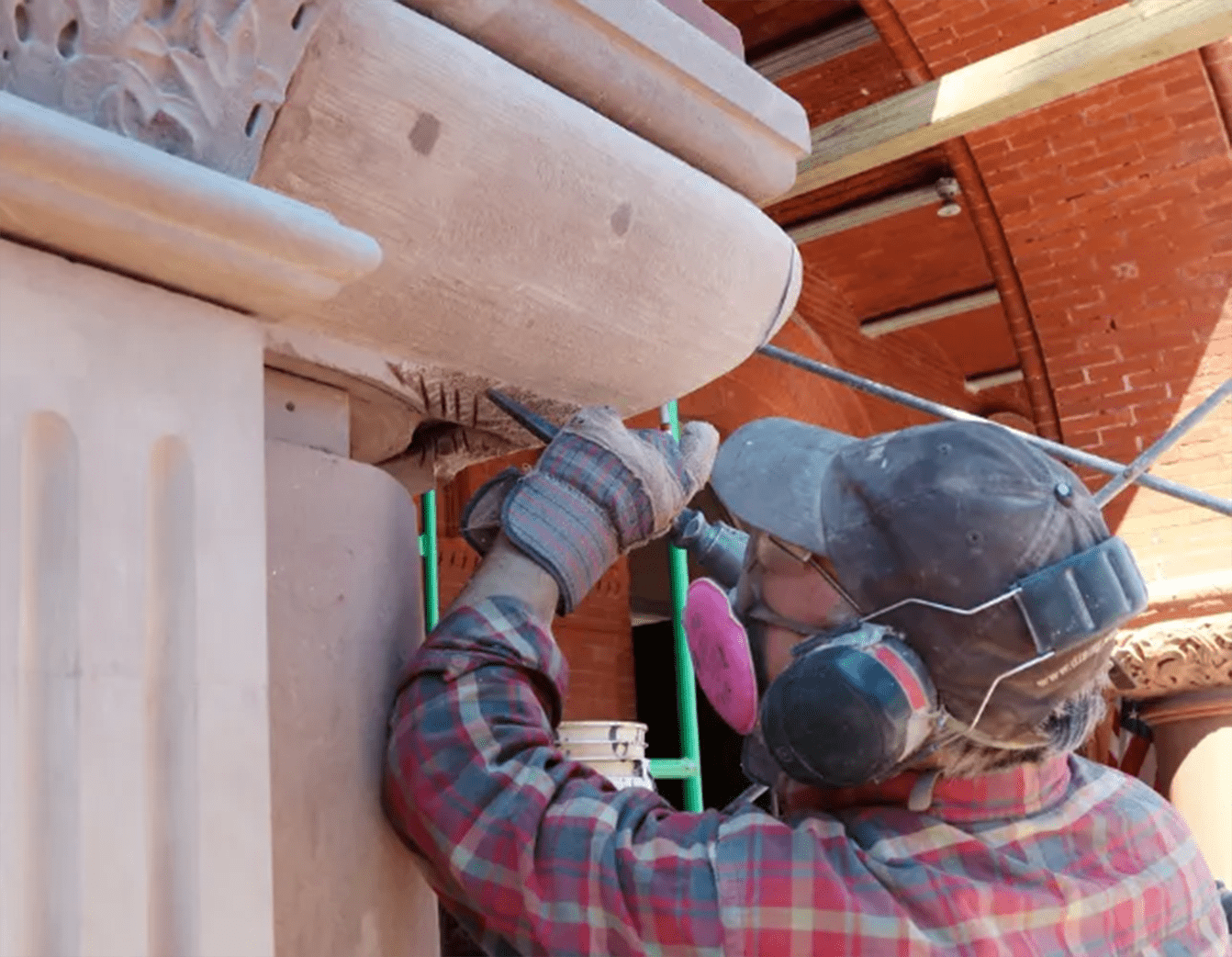 Worker repairing stone architectural detail, wearing protective gear, red brick backdrop.