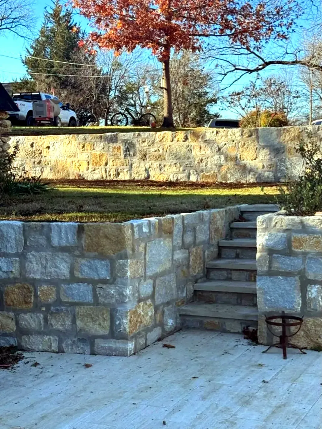 Stone retaining walls with stairs, cement patio, grass, and autumn tree in background.