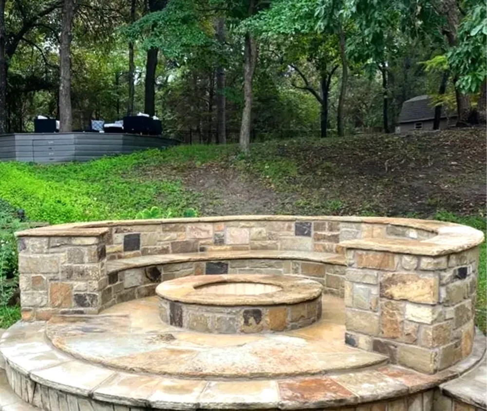 Stone fire pit with curved seating in a grassy outdoor area, surrounded by trees.