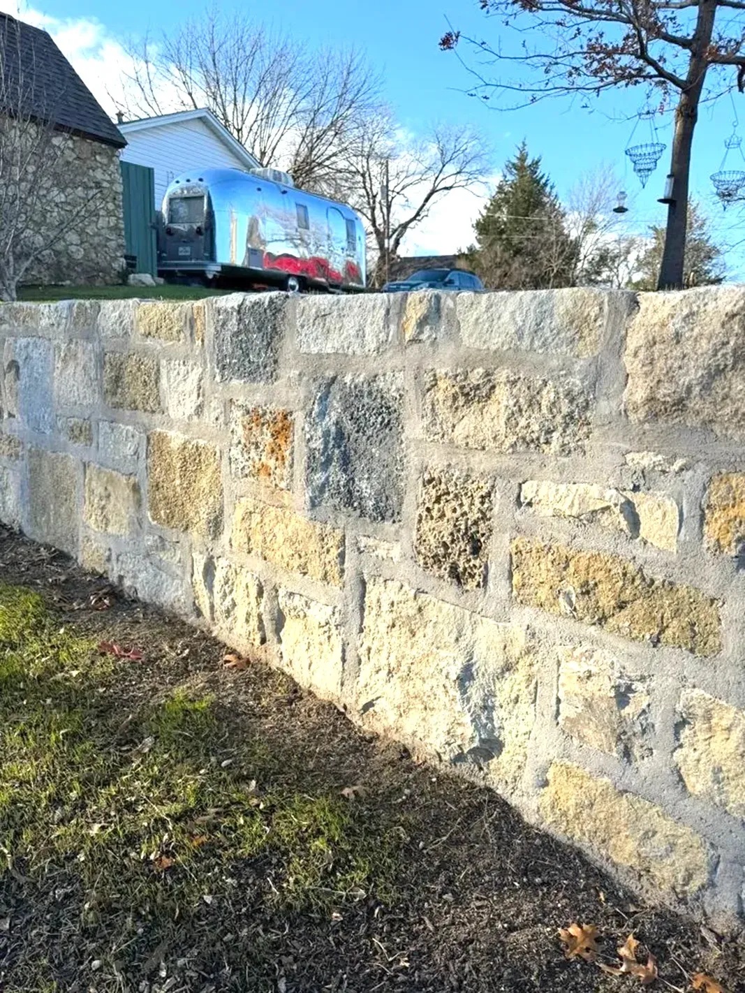 Stone wall in front of green grass. Airstream trailer behind wall. Blue sky and bare tree branches.