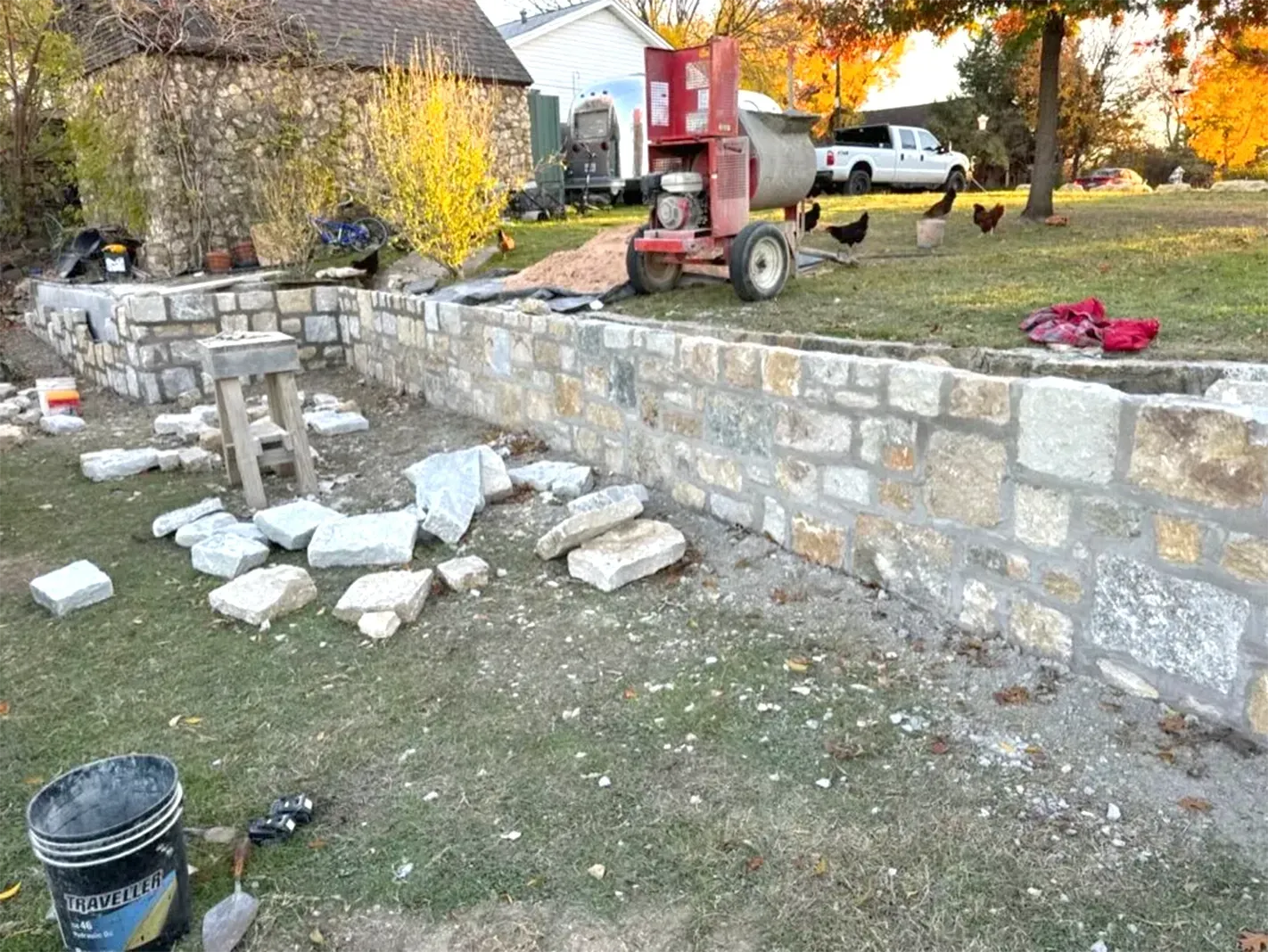 Stone retaining wall under construction in a yard; tools, mixer, and chickens visible.