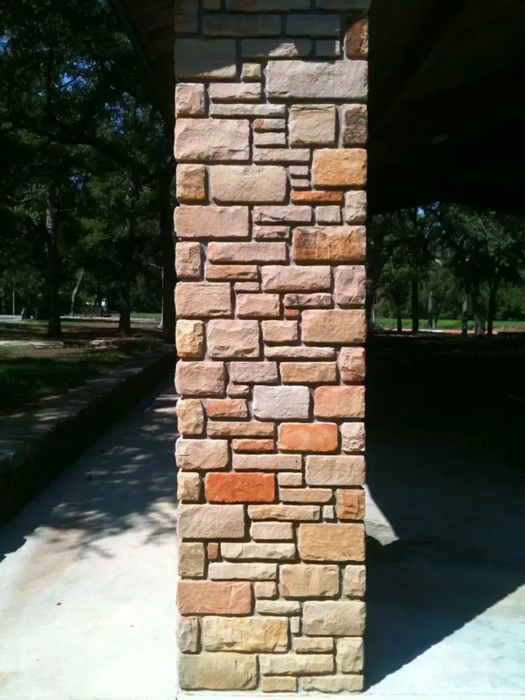 Stone-covered pillar, tan and brown stones, supporting a roof. Outdoors with trees and a pathway visible.
