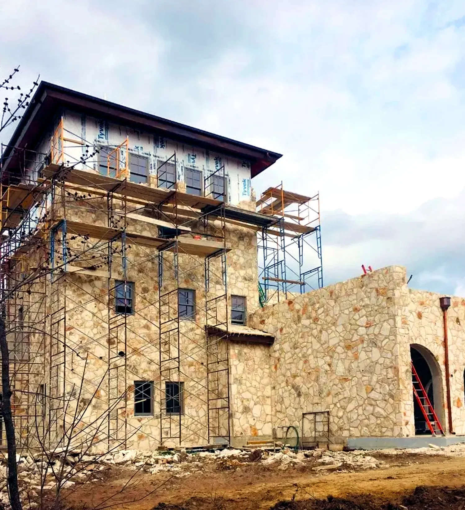 Stone building under construction with scaffolding, a red ladder, and blue sky.