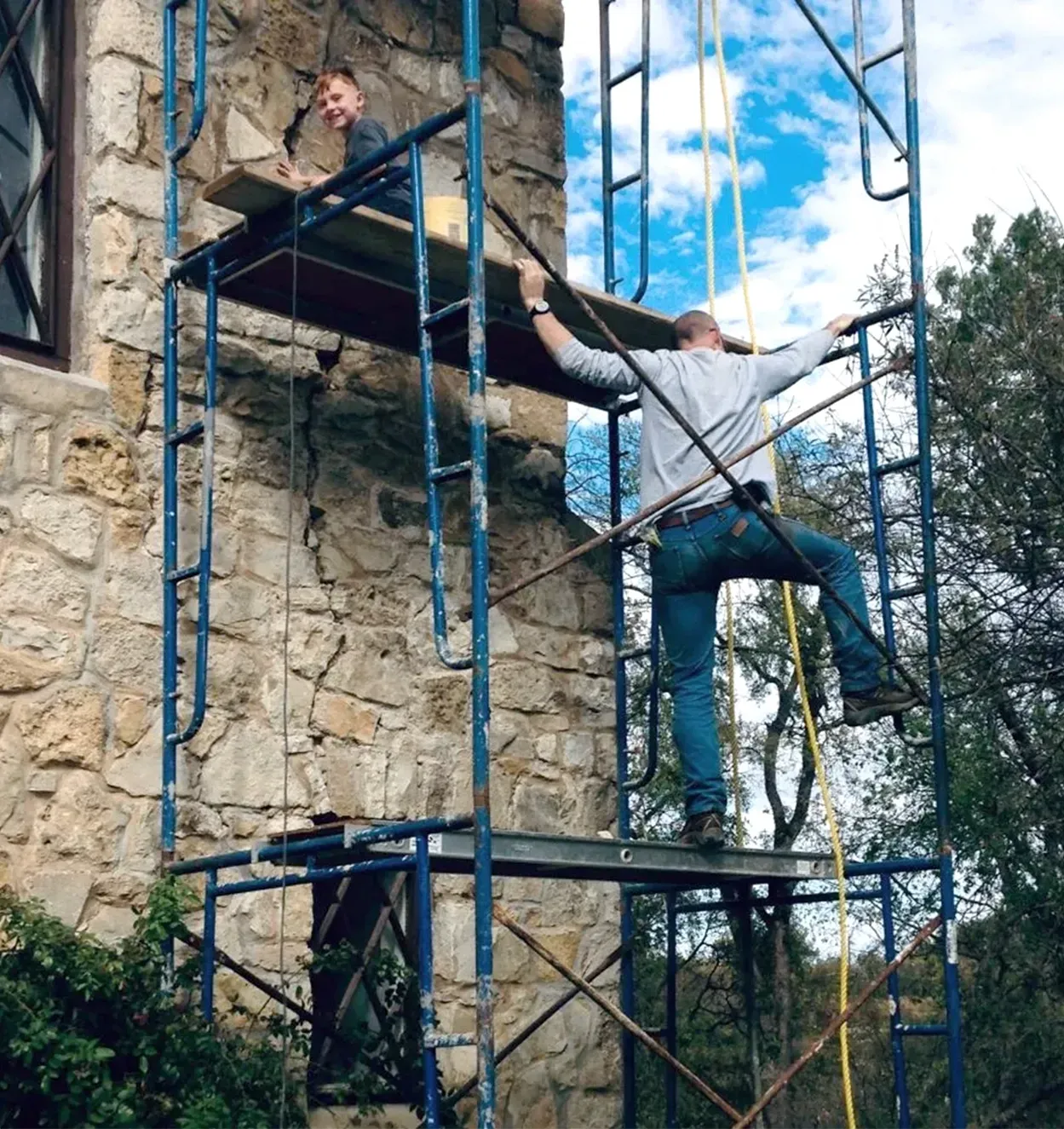 Two men on a scaffolding, working on a stone building exterior. One stands at the top, the other climbs.