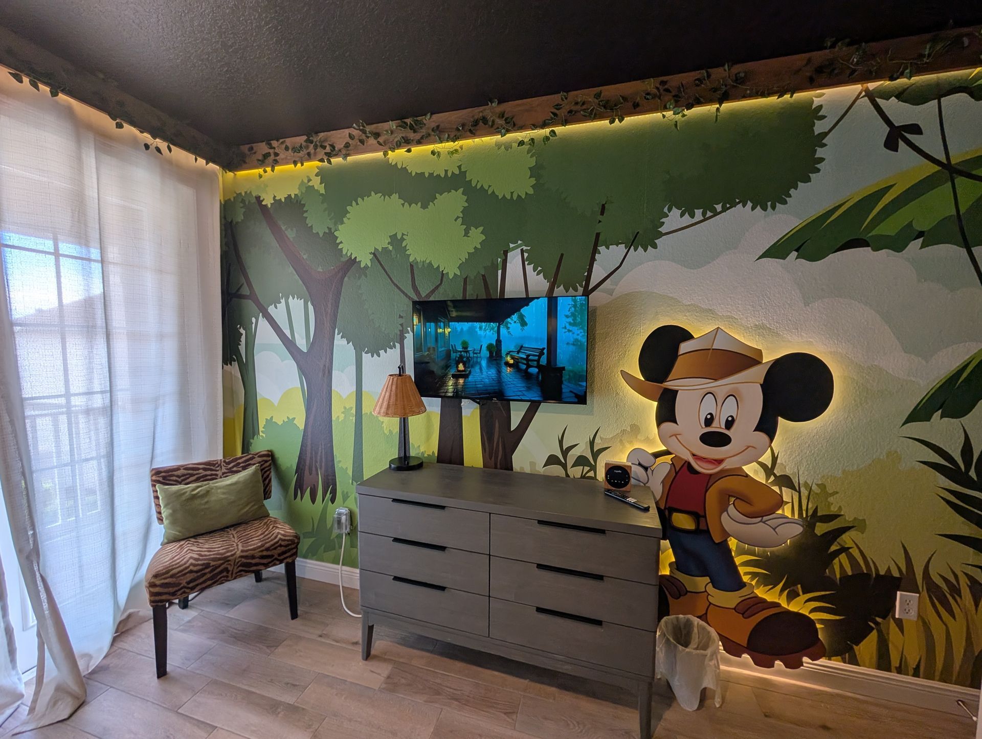 Jungle-themed nursery with mural, Mickey Mouse, TV, dresser, chair, and curtains. Green and brown colors.