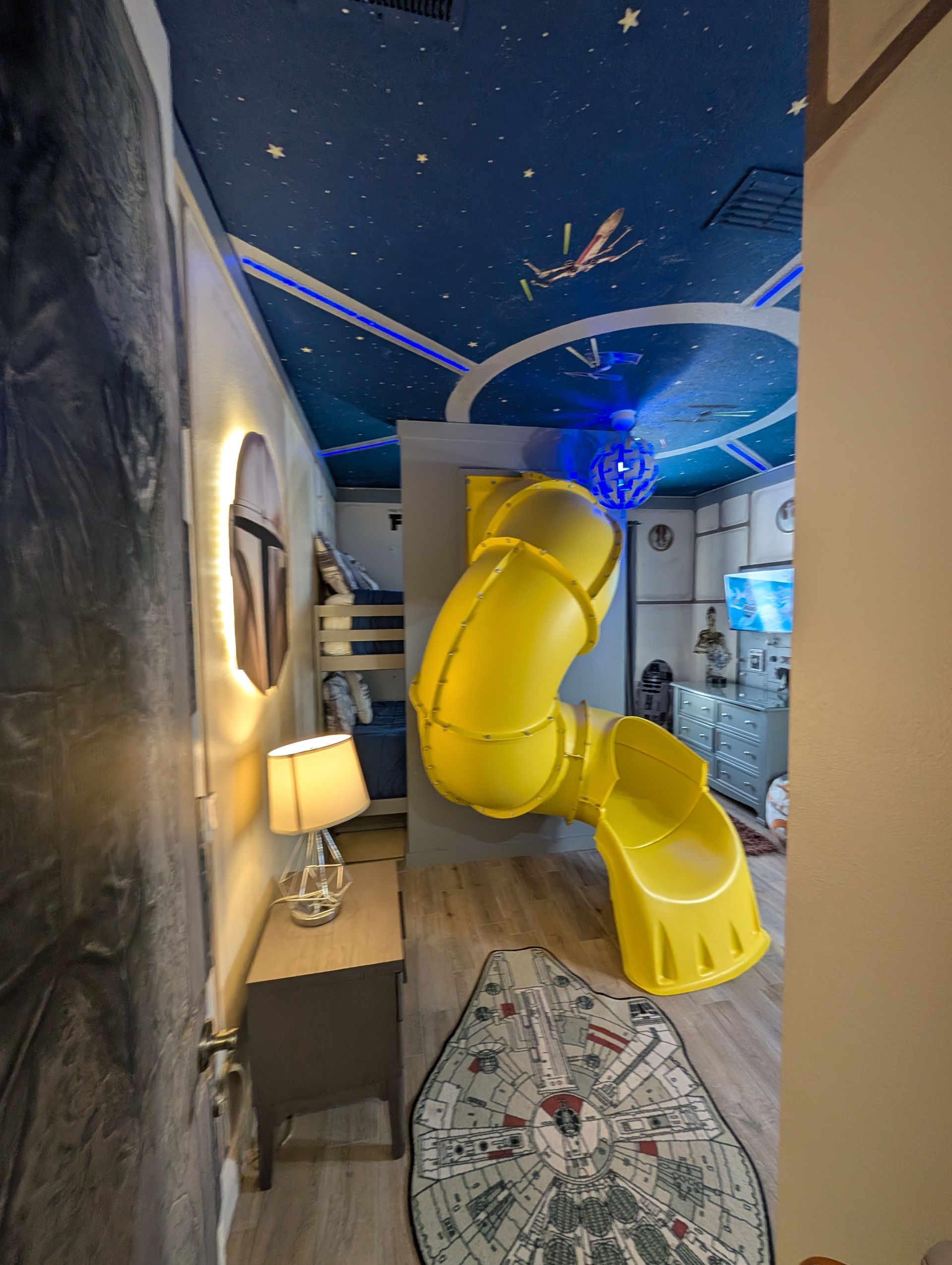 Star Wars themed bedroom with yellow slide. Blue starry ceiling, Millennium Falcon rug, nightstand, and bunk beds.