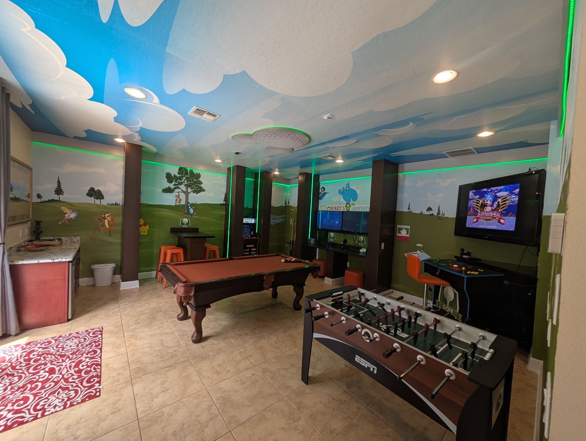 Game room with foosball, pool table, and TV, decorated with a sky mural and colorful lighting.