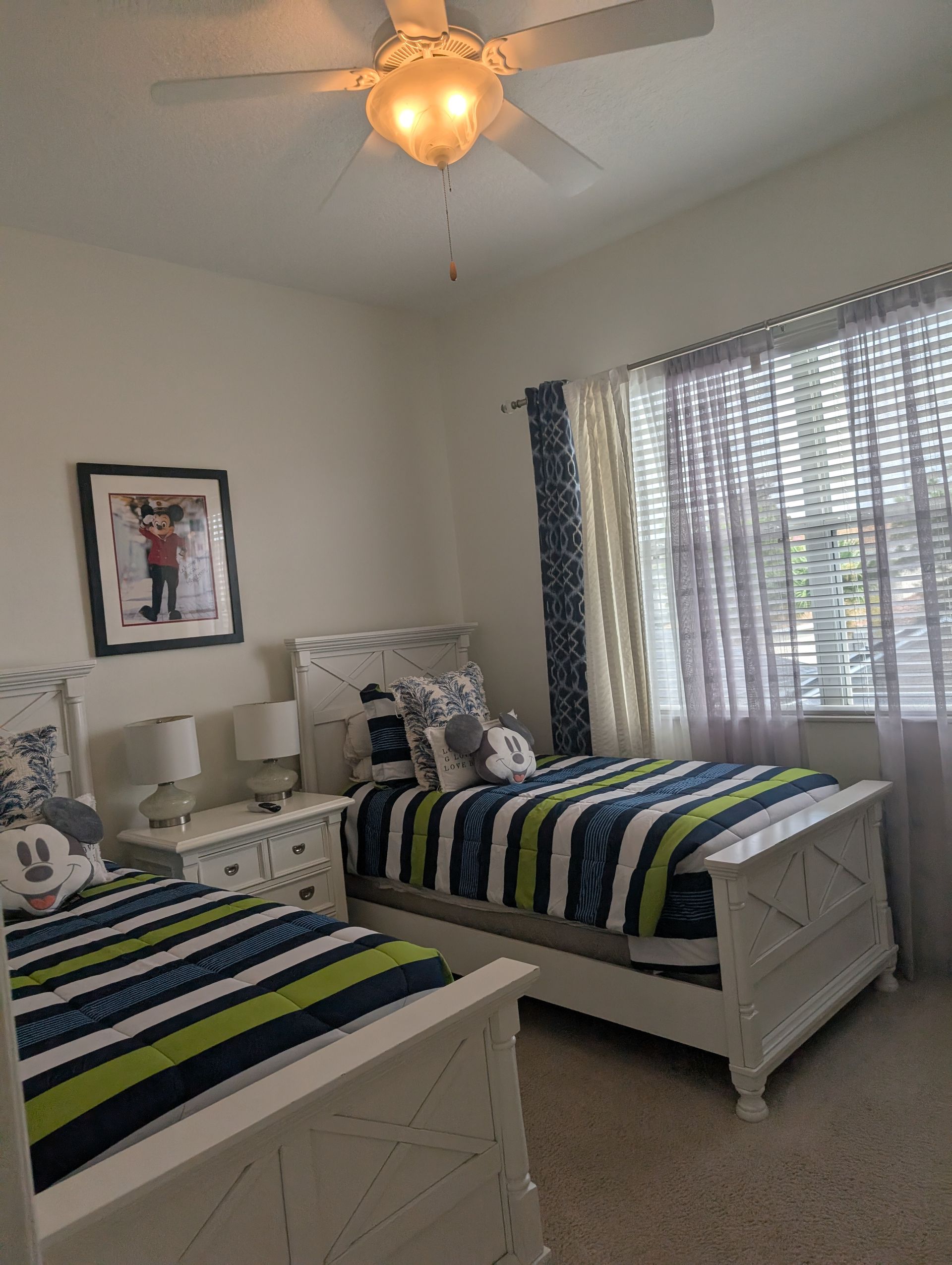 Two twin beds in a bedroom, white furniture, striped bedding, photo on wall, light curtains and ceiling fan.