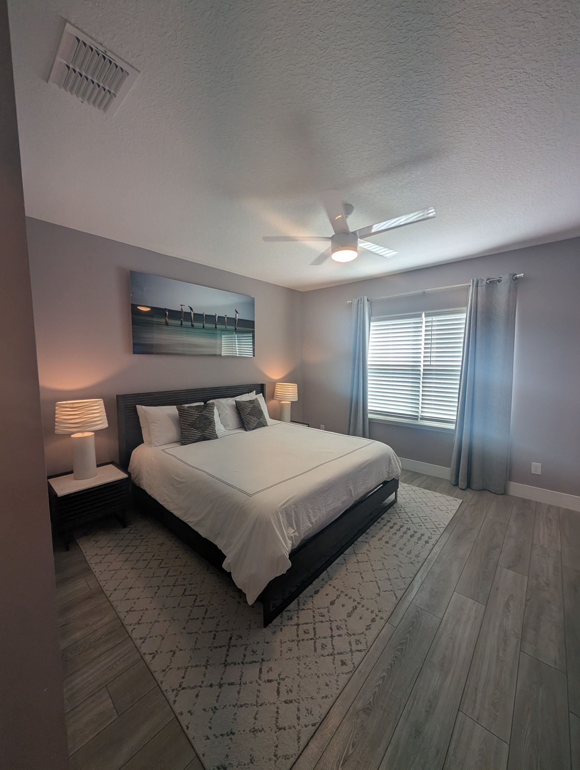 Bedroom with a bed, nightstands, art, a rug, and a window with blinds and curtains.