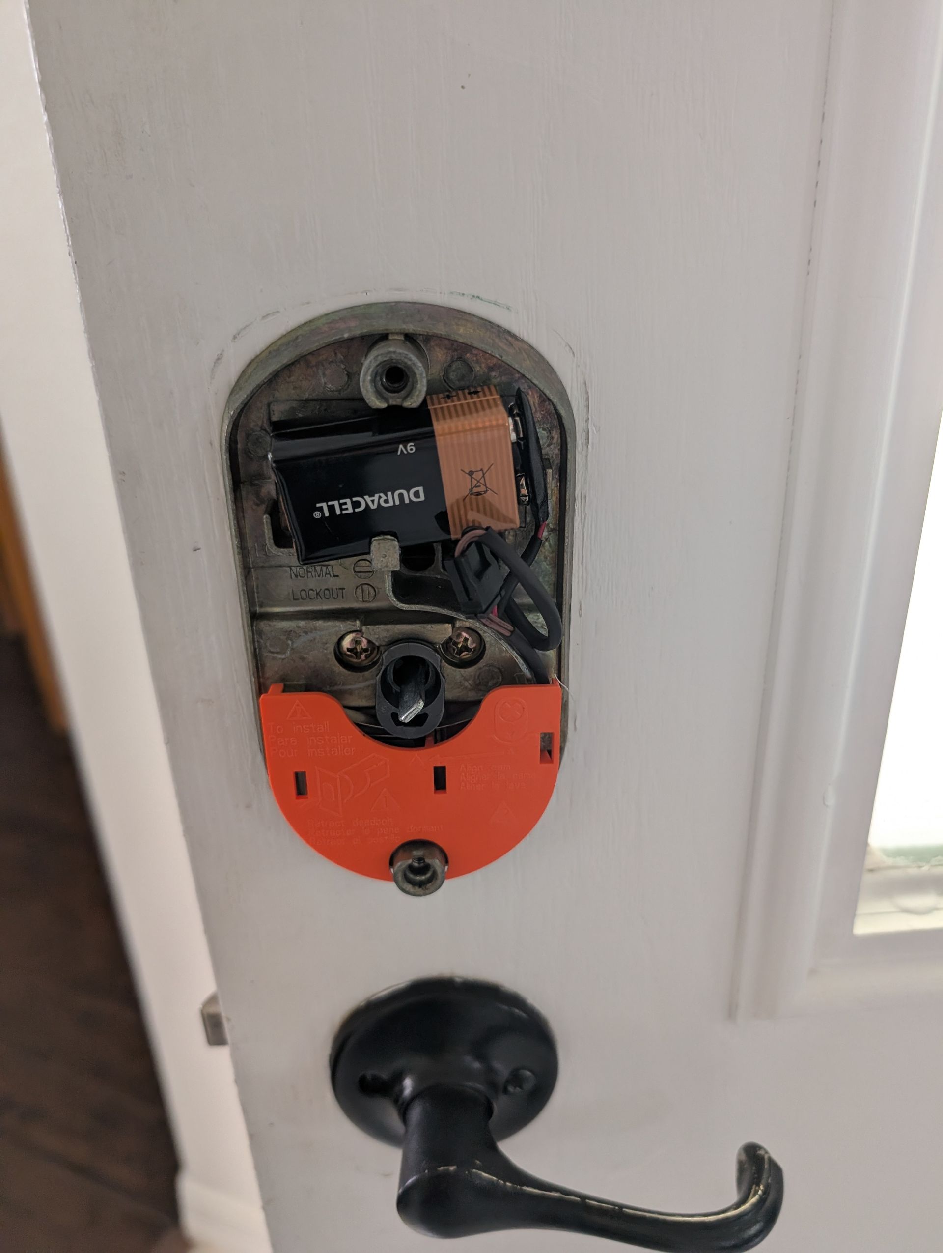 Smart lock mechanism, exposed on a white door; orange cover, battery, and black wires.