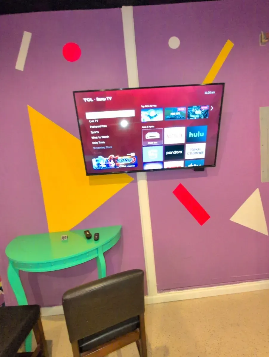 Purple wall with geometric shapes and a TV. A teal table and chair are in front of it.