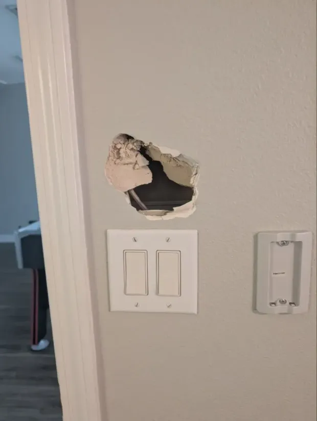 Hole in a light gray wall above two light switches and a thermostat mount, near a door frame.