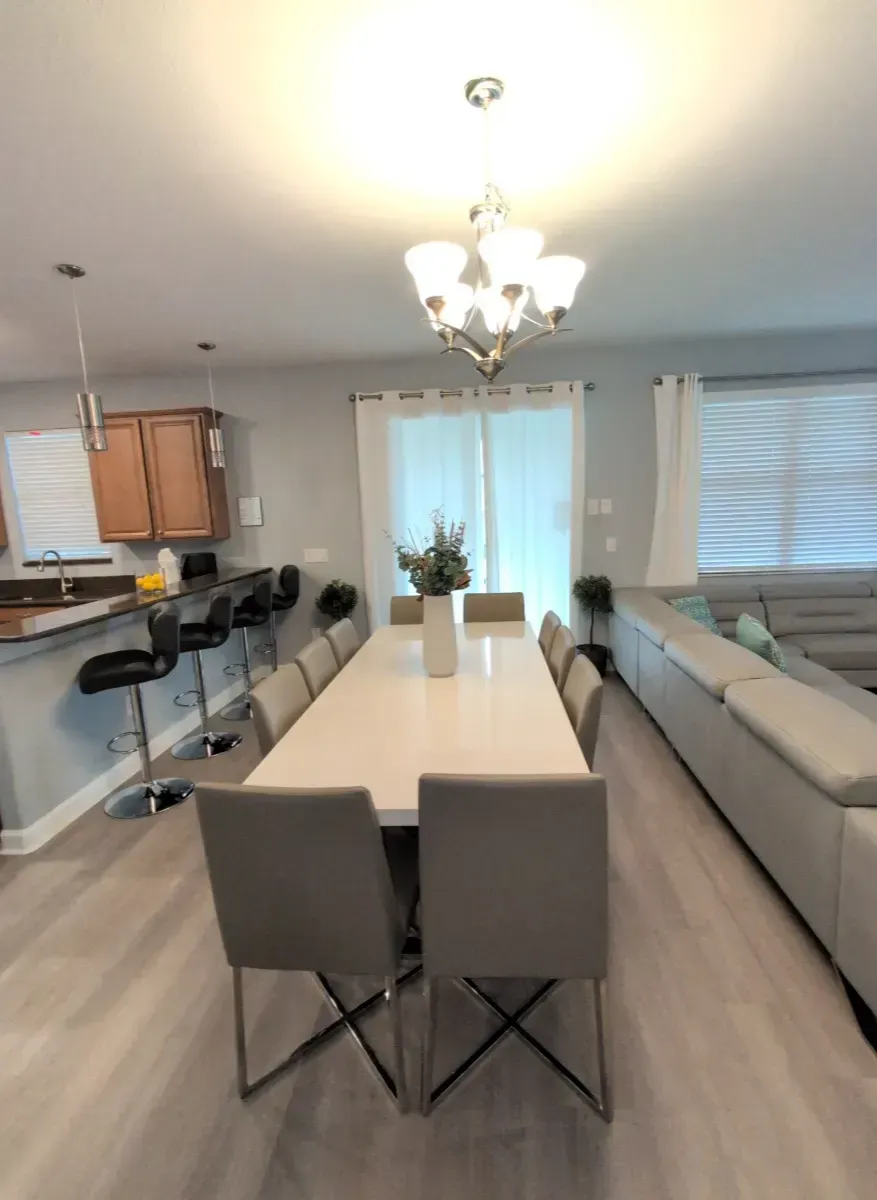 Spacious dining room with long white table, gray chairs, and a chandelier. Kitchen with bar stools and large sectional couch.