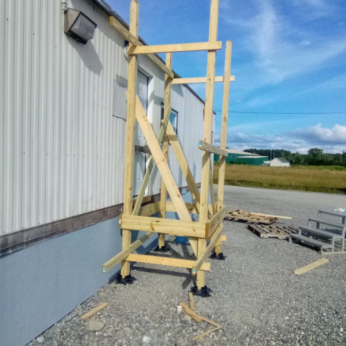 Wooden scaffolding structure built against a building, on a gravel surface. Blue sky visible.