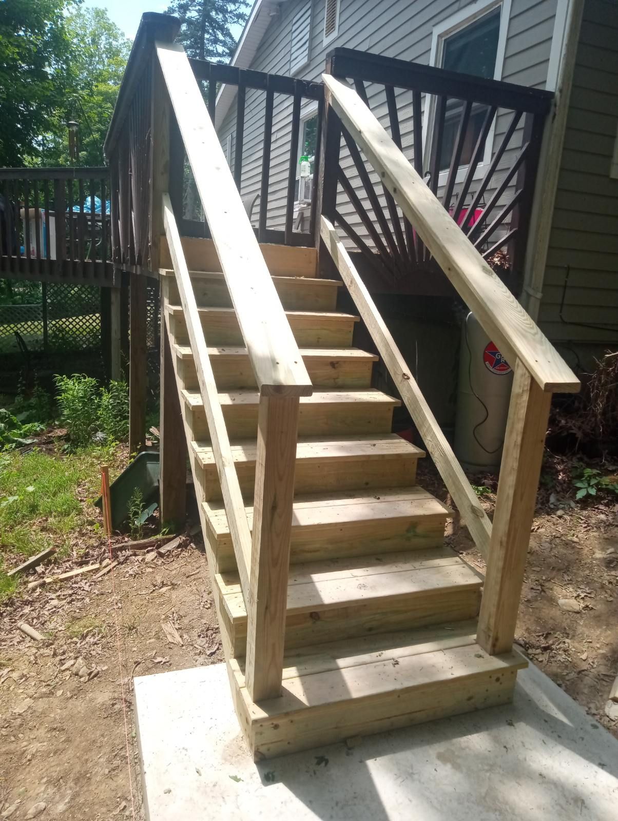 Wooden outdoor staircase leading to a deck, with handrails and concrete landing, set against a house and greenery.