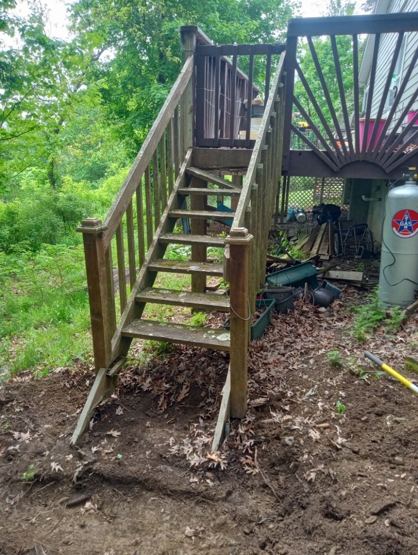 Wooden outdoor staircase leading up to a deck, set in a wooded area. Stairs show wear, dirt ground.