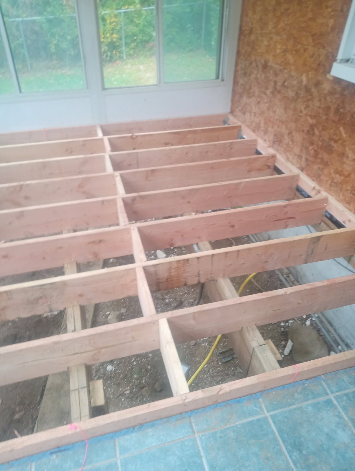 Wooden floor joists being constructed inside a room with windows and exposed plywood walls.