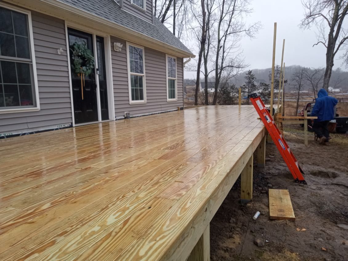 Newly built wooden deck on a house. Person in blue jacket. Red ladder. Overcast, wet conditions.