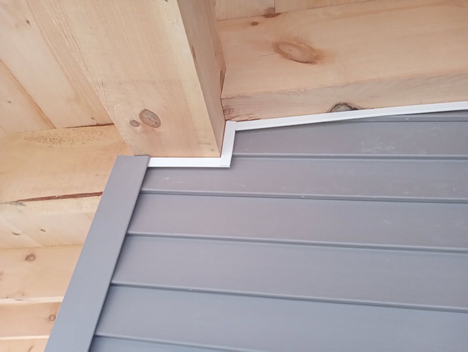Gray siding with white trim meeting a wooden beam in an outdoor setting.