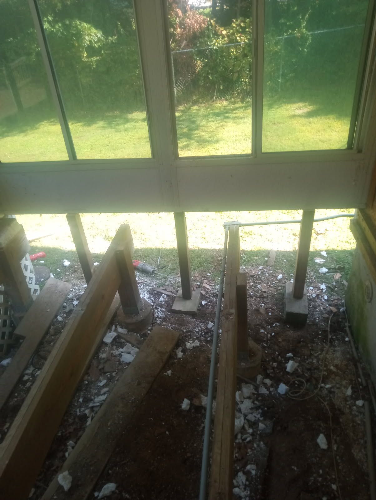 Sunroom under construction: Interior view with temporary support jacks, wood debris, and outdoor view through windows.