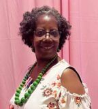 A woman wearing glasses and a green necklace is standing in front of a pink curtain.
