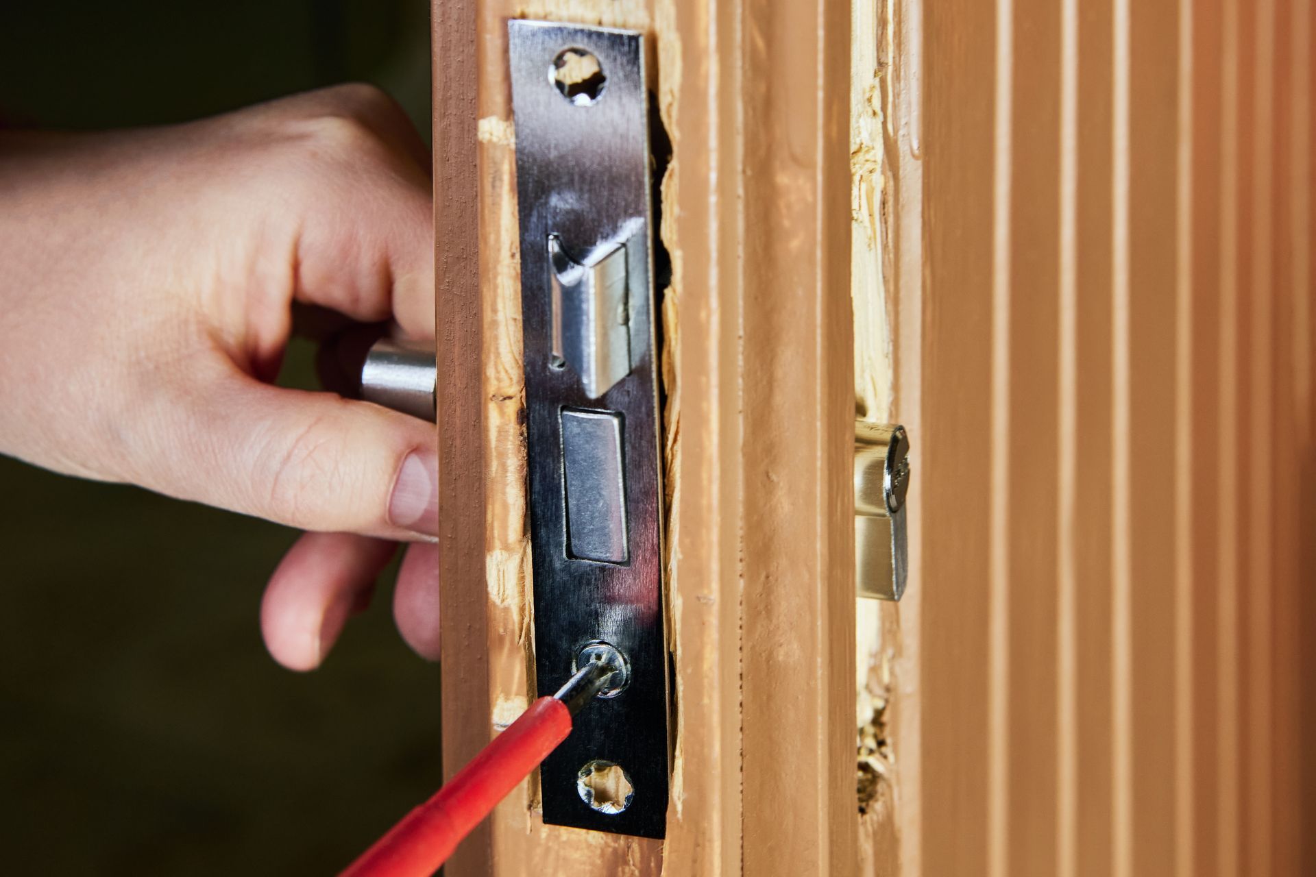Hand holding a red screwdriver near a door latch on a wooden door frame